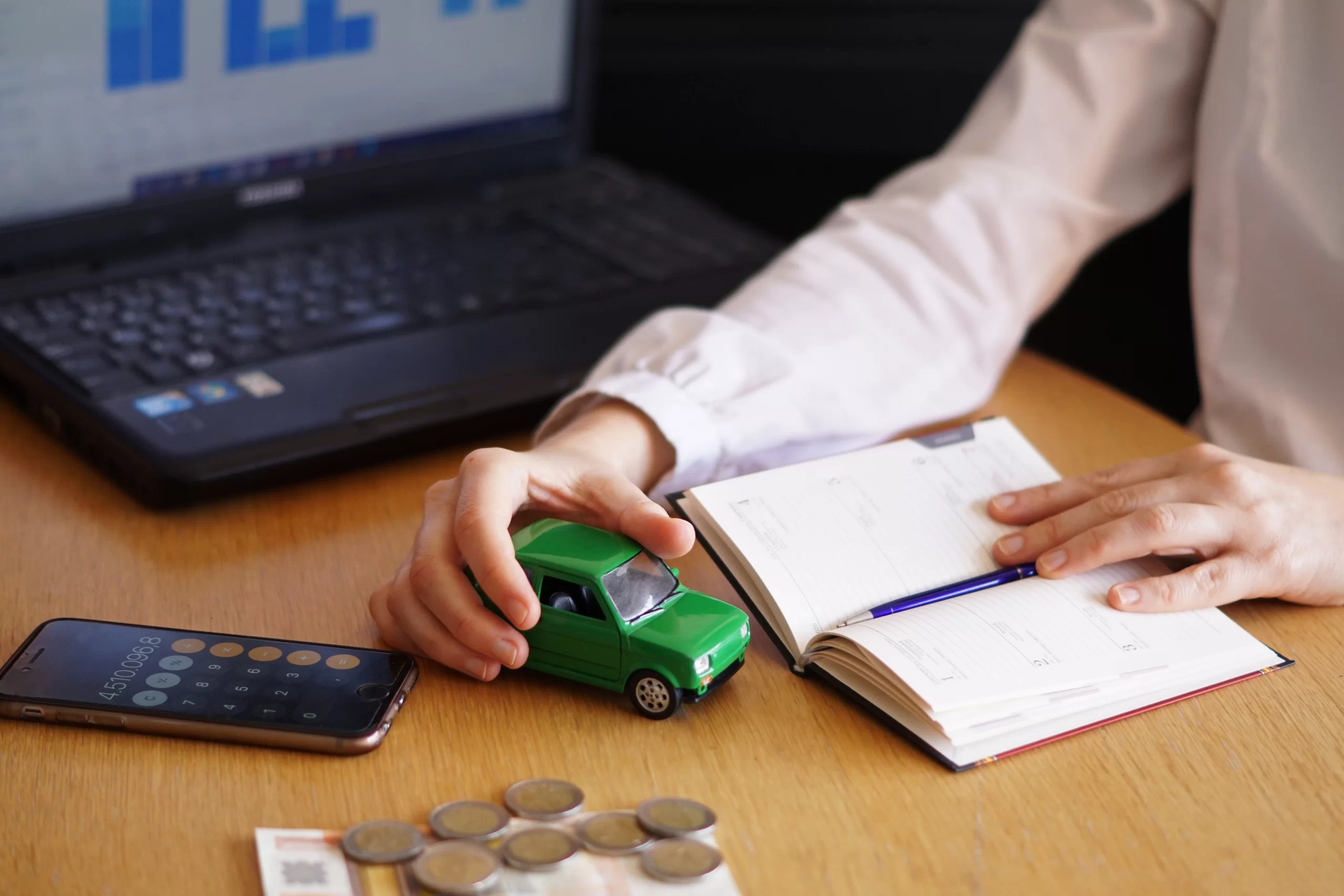 Imagem com um homem segurando um carrinho de brinquedo em uma mesa de escritória e mais agenda, moedas e calculadora e computador na mesa