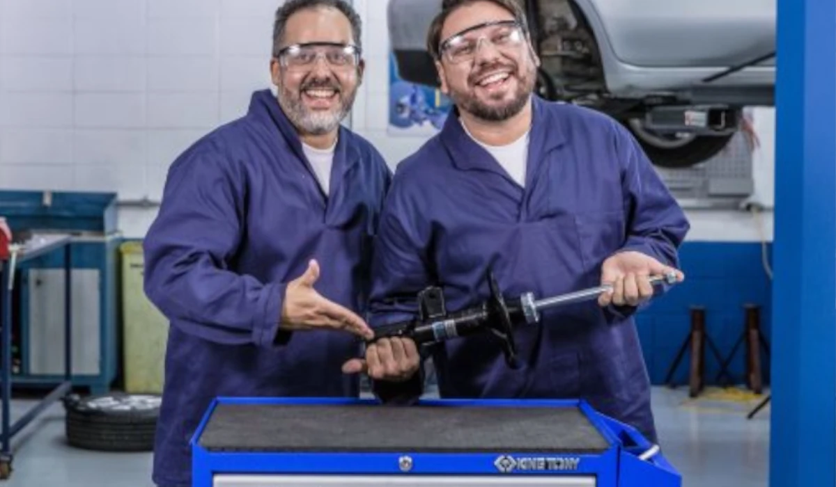 Imagem com dois mecânicos no centro fazendo pose com amortecedor nas mãos de um deles